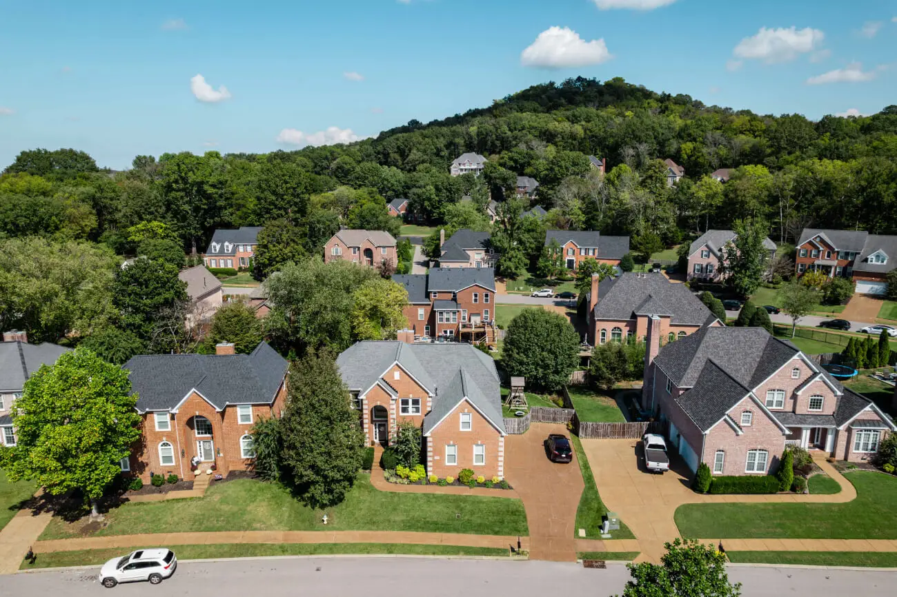 Aerial view of a suburban neighborhood with brick houses, green lawns, and tree-lined streets nestled near a wooded hill under a blue sky with scattered clouds. | Red Beard Roofing