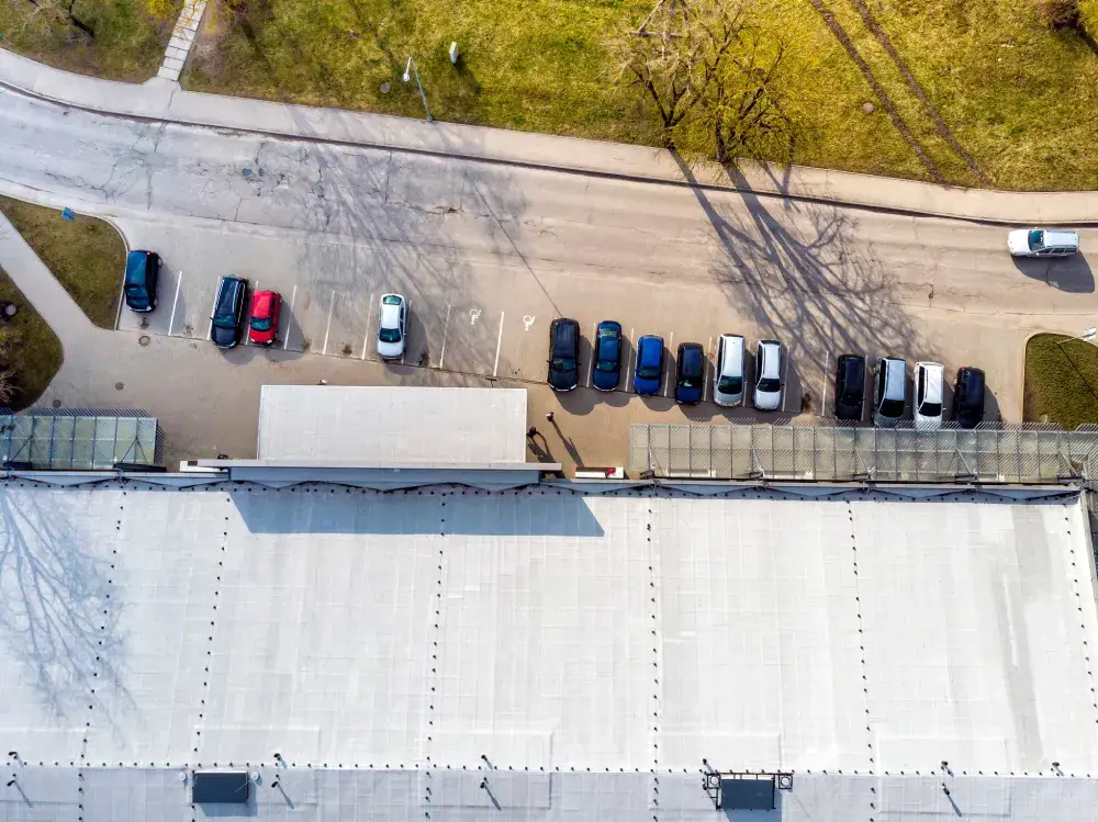 Aerial view of cars parked in a row beside a large commercial roof with a white surface. Shadows from trees fall across the ground and roof, while a grassy area appears at the top. Perfect for showcasing commercial roof repair projects. | Red Beard Roofing