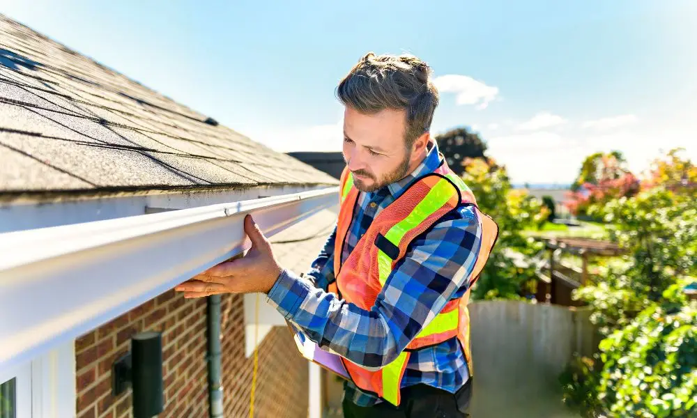 A professional contractor wearing a safety vest inspecting a home's gutters during a routine roofing and maintenance check on a residential building.