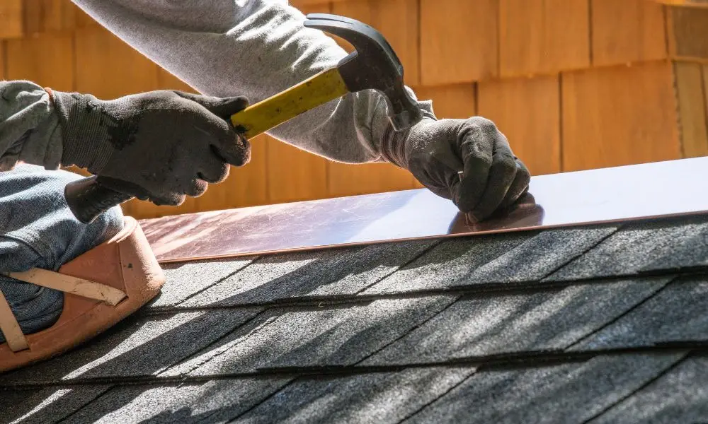 A roofer installation process showing a worker using a hammer to secure a copper roof flashing strip along the edge of an asphalt shingle roof.