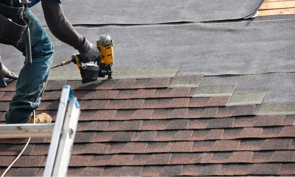 A roofer installing new shingles during a roof replacement project, using a yellow pneumatic nail gun to secure the asphalt shingles onto the underlayment.